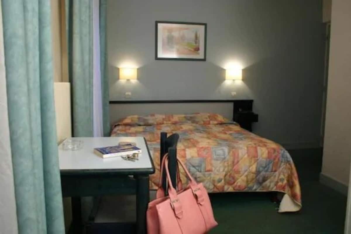 A hotel room with a bed, a bedside table with a lamp, a chair, and a table with a book and a handbag.