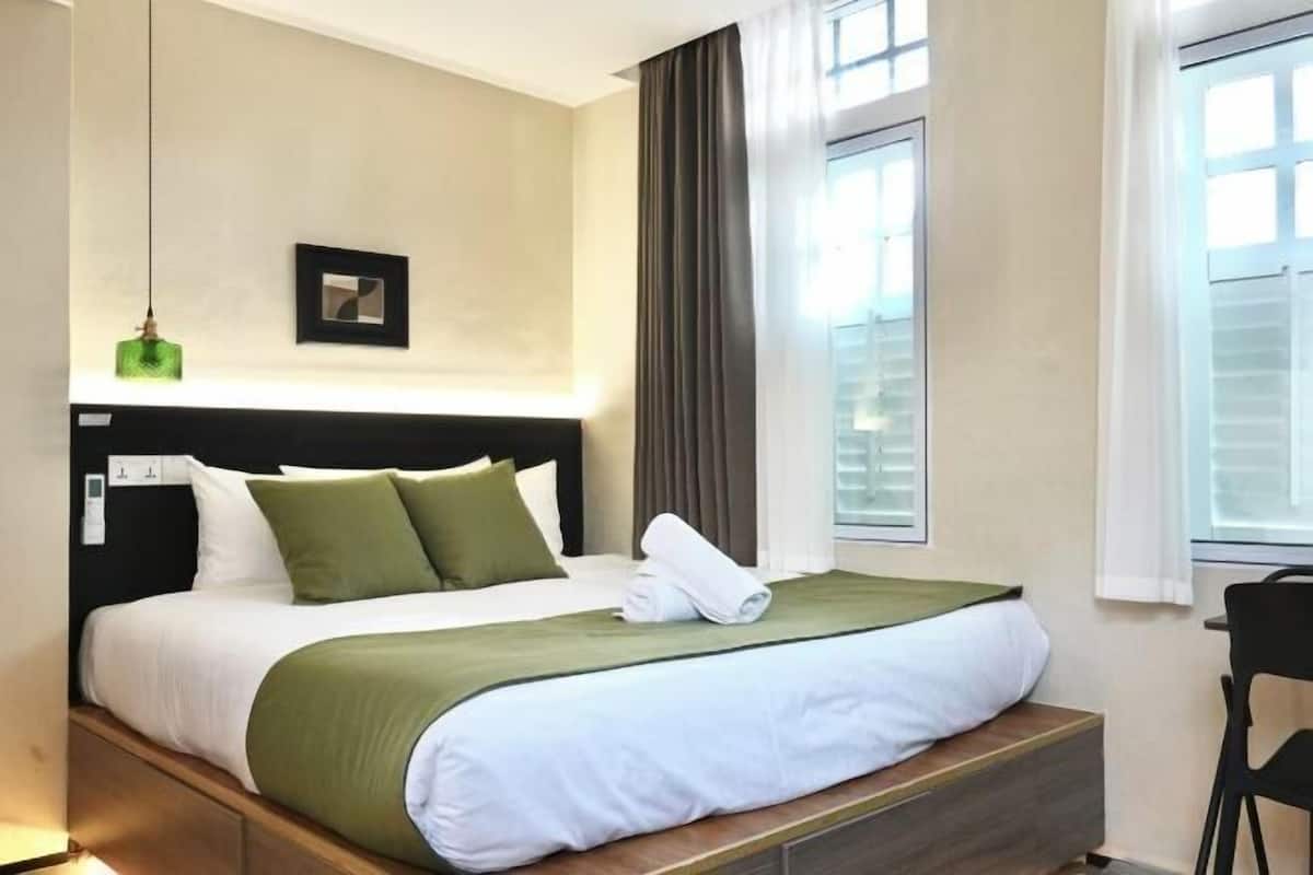 Quarto de hotel com cama, mesa de cabeceira, cadeira, janela com cortinas e um quadro na parede.