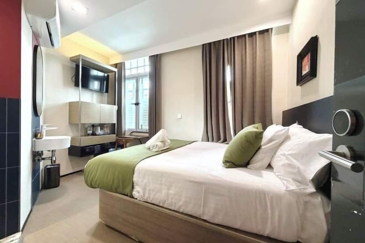 Quarto de hotel com cama, televisão na parede, secretária com cadeira e janela com cortinas.