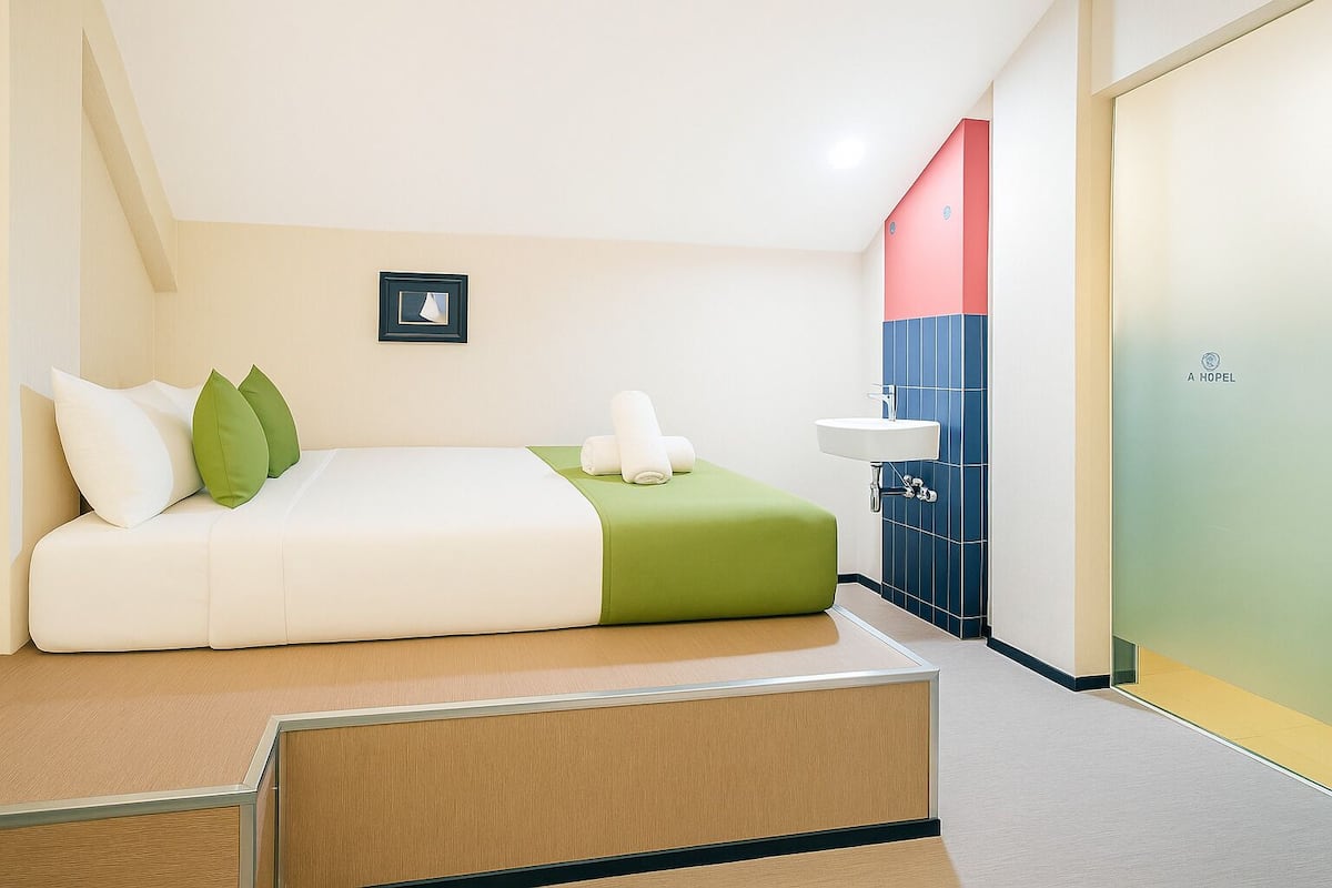Quarto de hotel moderno com cama, almofadas verdes, mesa de cabeceira, pia e porta com um logotipo.