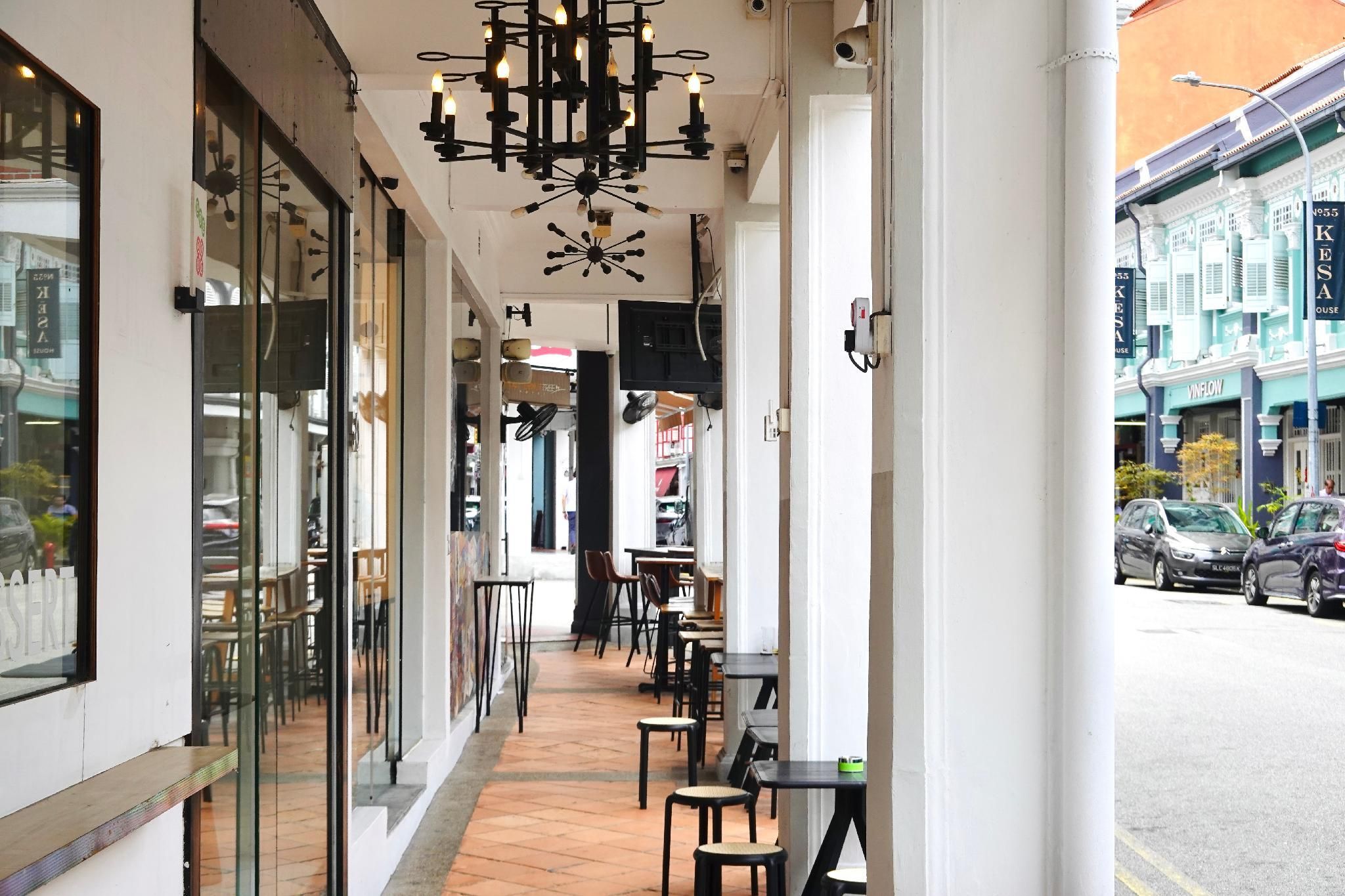 Un área de café al aire libre con mesas y sillas, un candelabro moderno y vista a la calle.