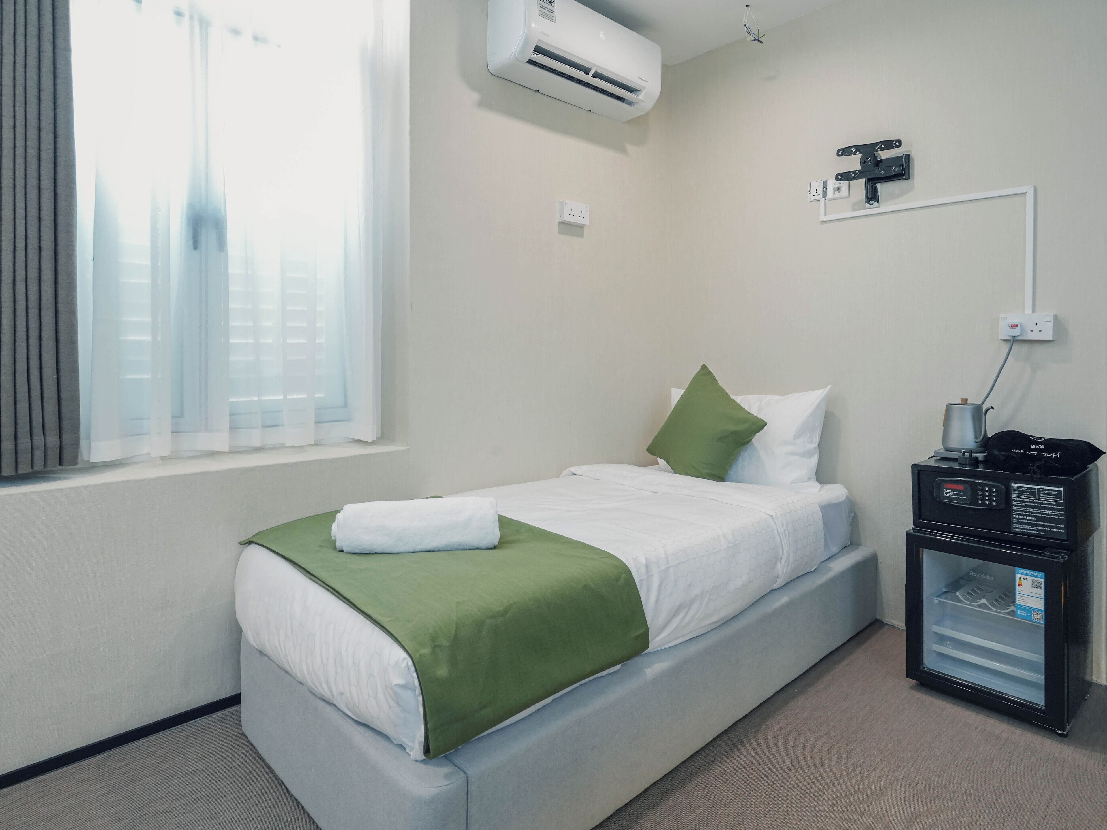 Ein Hotelzimmer mit einem Bett, einem grünen Kopfkissen, einer weißen Decke, einer Klimaanlage, einem kleinen Tisch mit einem Wasserkocher und einer Minibar.