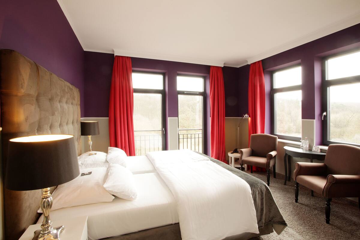 A hotel room with a large bed, two chairs, a small table, and a window with red curtains.