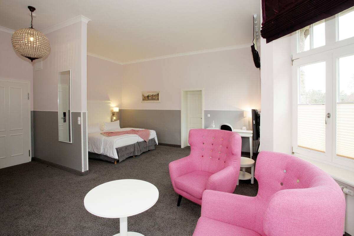 A hotel room with a bed, a pink armchair, a white table, and a chandelier.