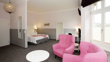 Une chambre d’hôtel avec un lit, un fauteuil rose, une table blanche et un lustre.