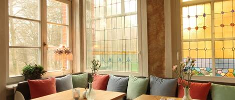 A cozy dining area with wooden tables and benches, large windows with stained glass, and colorful cushions.