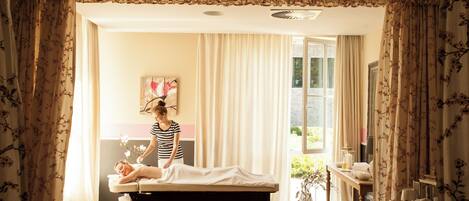 A room with a massage table, a person attending to a client, and a window with curtains.