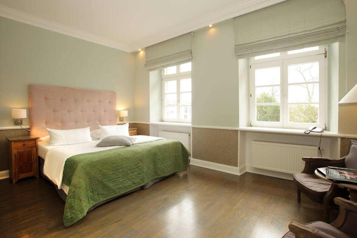 A bedroom with a large bed, bedside lamps, a window with blinds, a radiator, and a chair with a magazine.