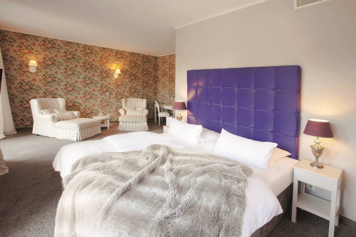 A hotel room with a large bed, a purple headboard, two armchairs, a small table, and floral wallpaper.