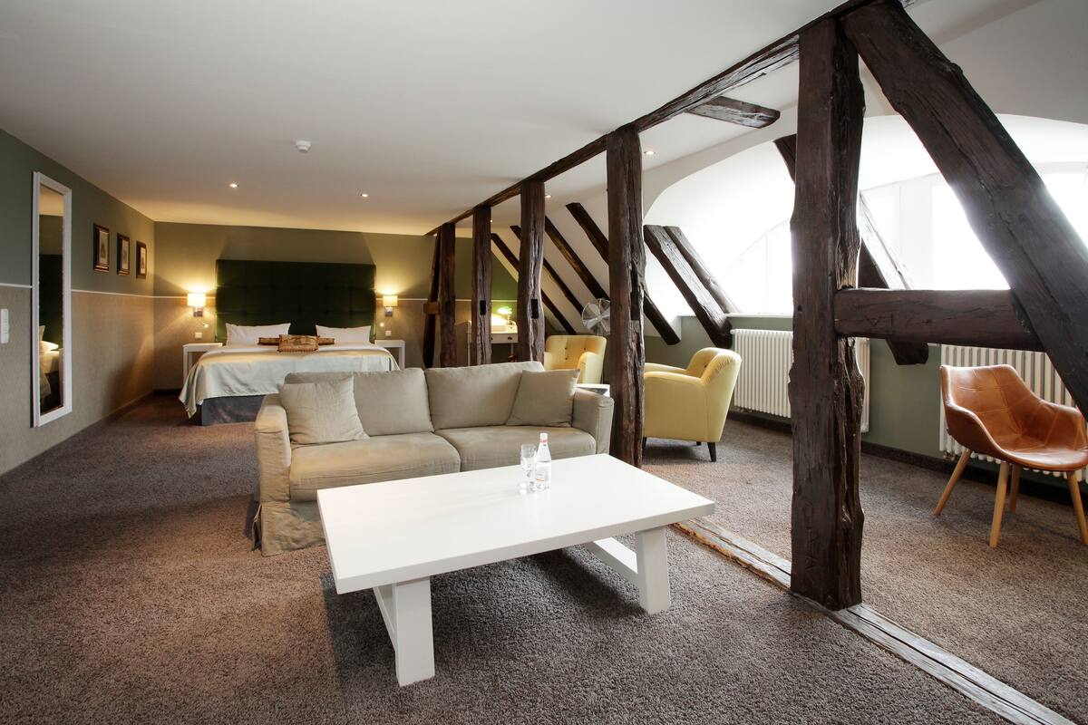 A room with a bed, a sofa, a coffee table, and a chair. There are wooden beams on the ceiling and a radiator on the wall.