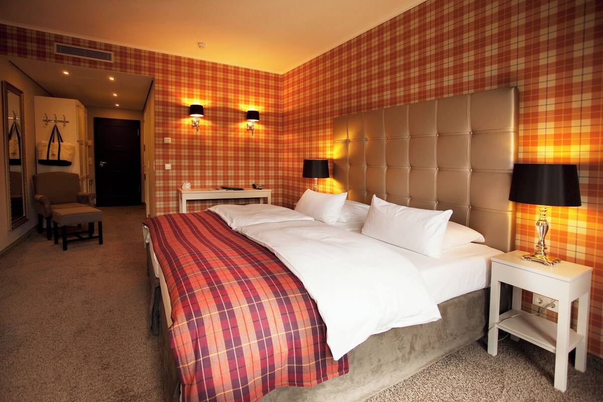 A hotel room with a large bed, two bedside tables with lamps, a headboard, a bench, and a wardrobe.
