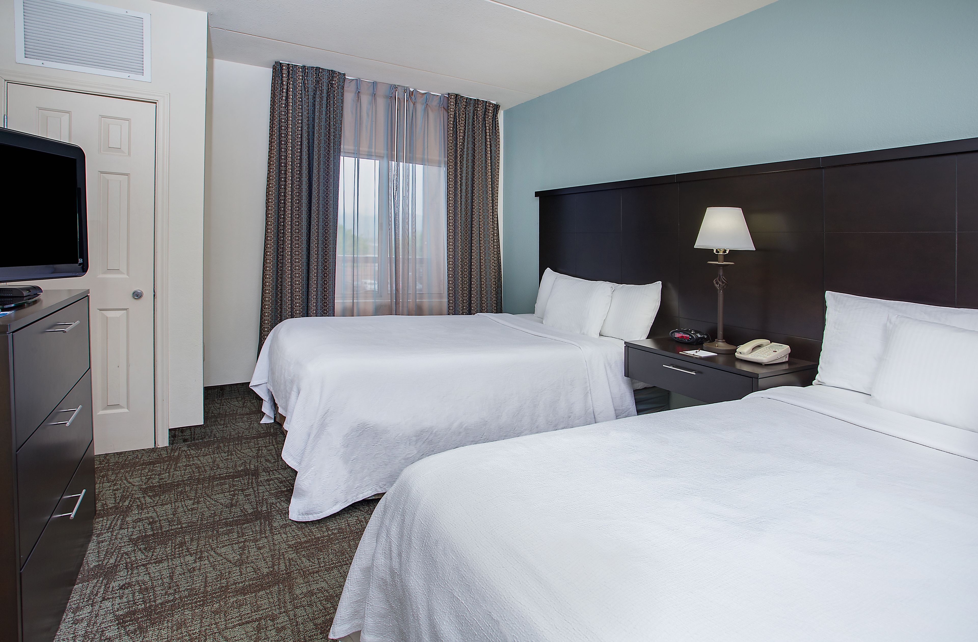 Quarto de hotel com duas camas, uma mesa com luminária, telefone e televisão em um aparador.