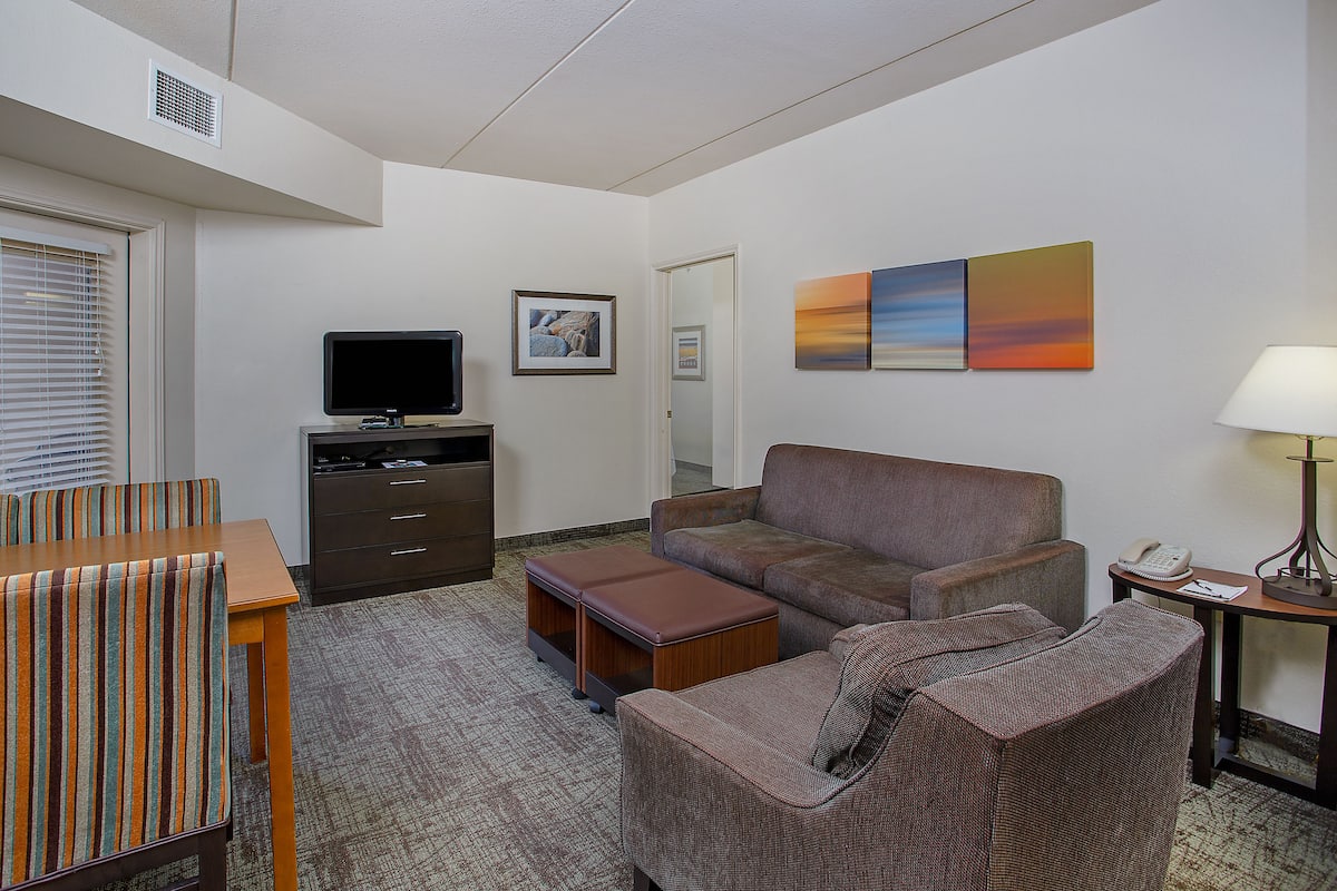 Una habitación de hotel con sofá, sillón, mesa de madera, televisor y una obra de arte en la pared.