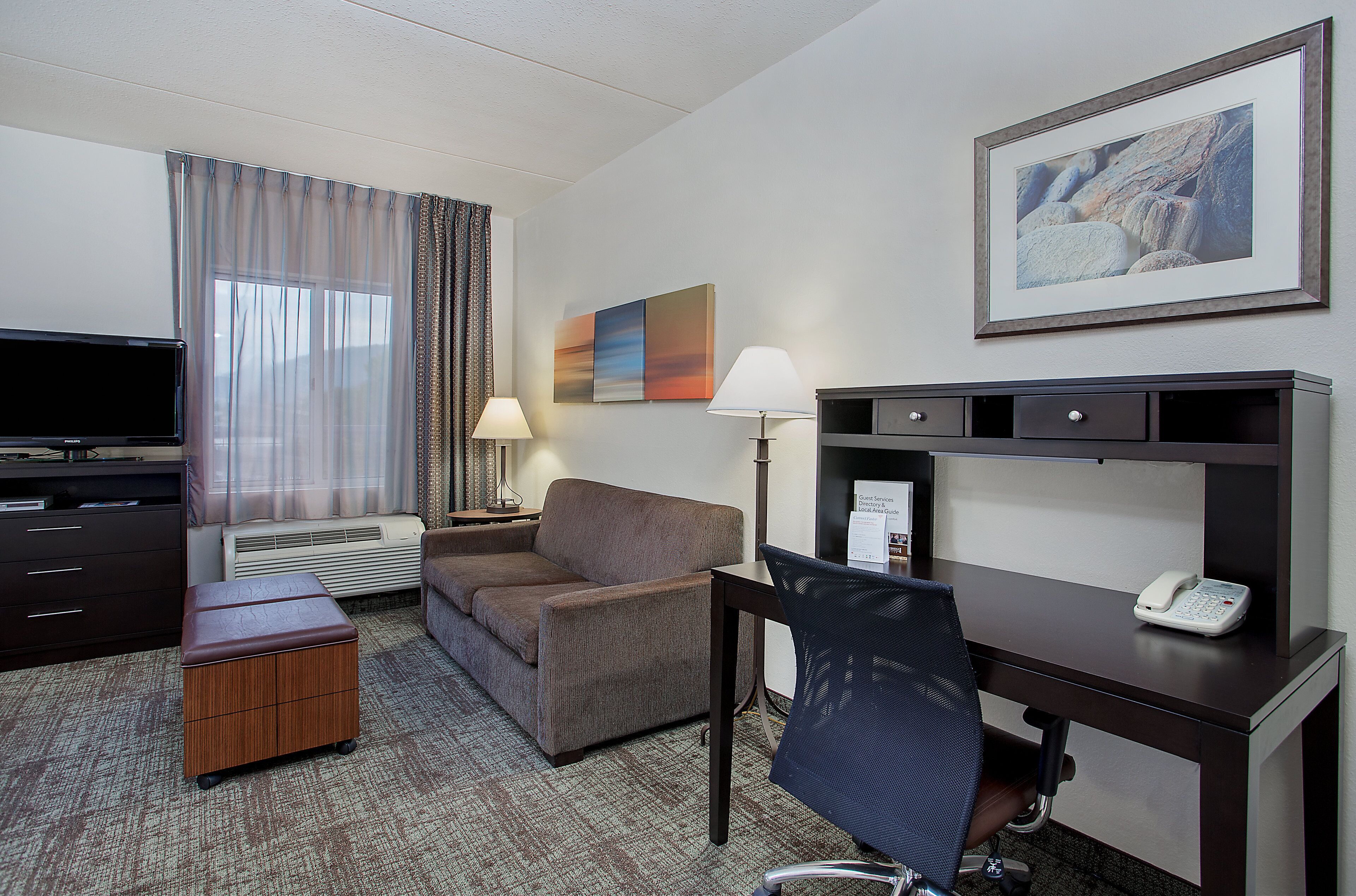 A hotel room with a flat-screen TV, a sofa, a desk with a phone, a chair, a lamp, and a painting on the wall.