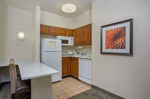 A compact kitchen with wooden cabinets, a white refrigerator, a microwave, and a dishwasher. A framed floral artwork is on the wall.