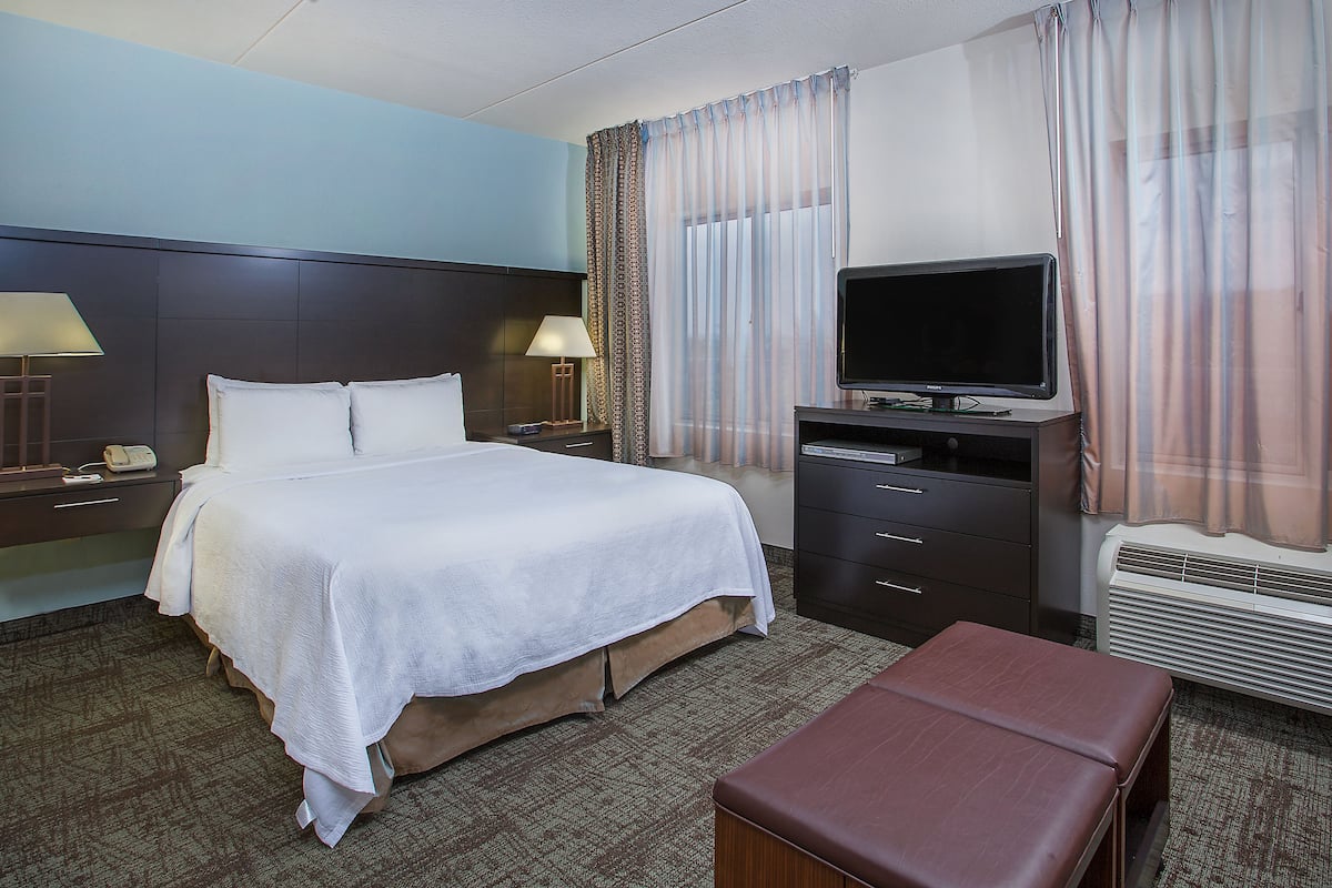 Una habitación de hotel con una cama, lámparas de noche, un televisor, un escritorio con una computadora y un banco.