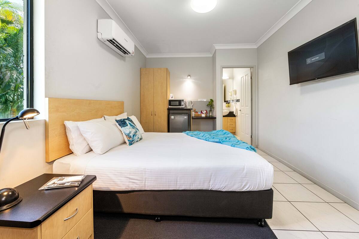 A hotel room with a bed, bedside table, lamp, air conditioning unit, TV, and a view of the kitchen area.