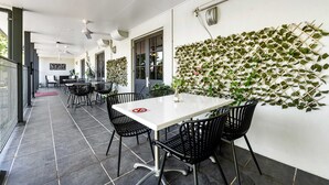 Un área de estar al aire libre techada con una mesa blanca y sillas negras, una pared cubierta de plantas trepadoras y un piso de baldosas.