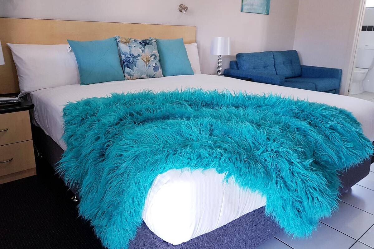A neatly made bed with a fluffy turquoise blanket, a wooden headboard, and a blue sofa in a modern room with a tiled floor.