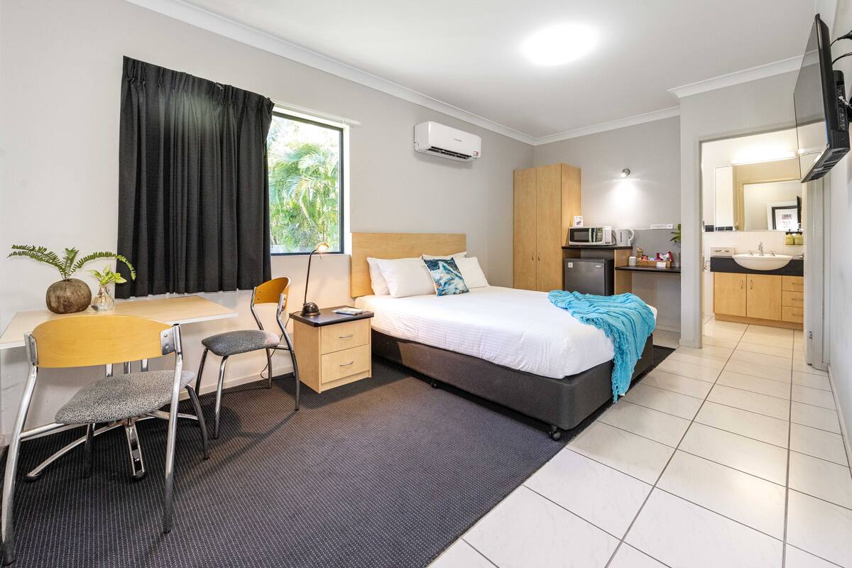 A modern hotel room with a bed, desk, chair, and kitchenette.