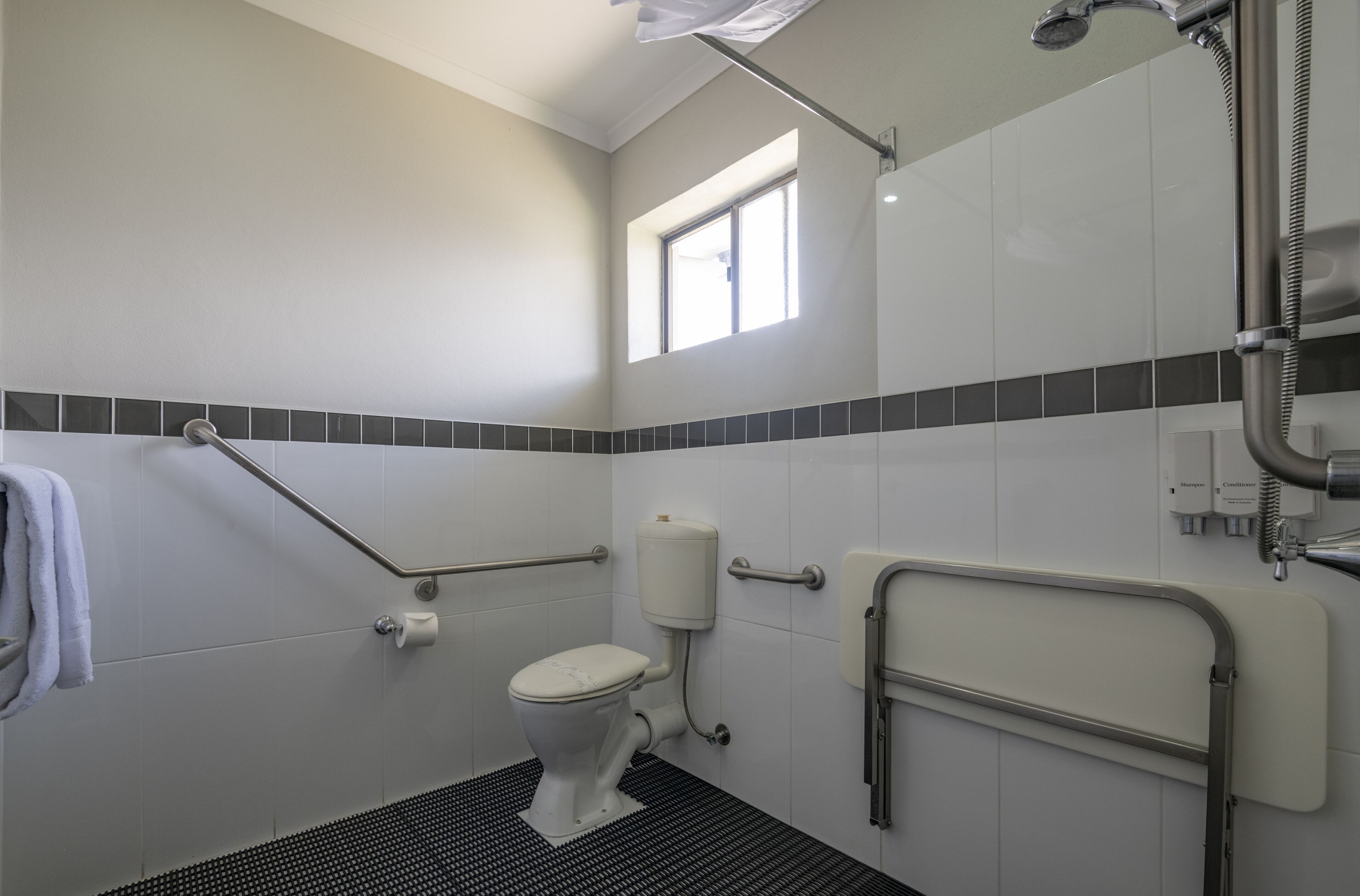 Une salle de bain équipée de toilettes, de barres d’appui, d’une douche et d’une fenêtre.