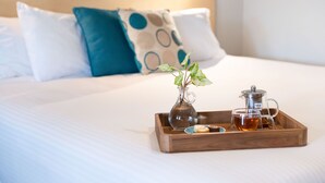 Una cama bien tendida con una bandeja que sostiene un jarrón de cristal con una planta, un calentador de agua y una taza de té.