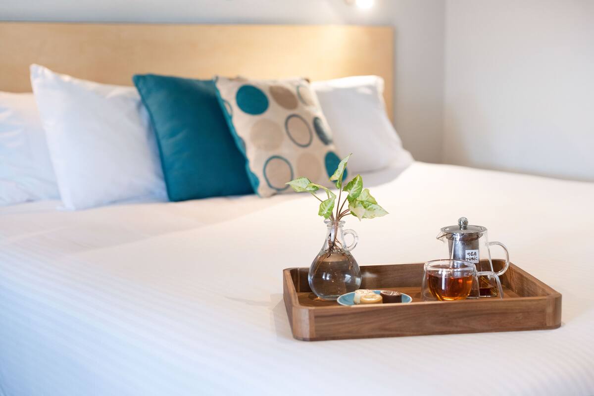 A neatly made bed with a tray holding a glass vase with a plant, a teapot, and a cup of tea.