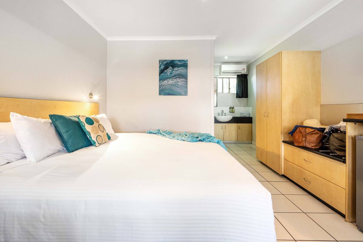 A hotel room with a large bed, a wooden wardrobe, a bathroom with a sink and mirror, and a painting on the wall.