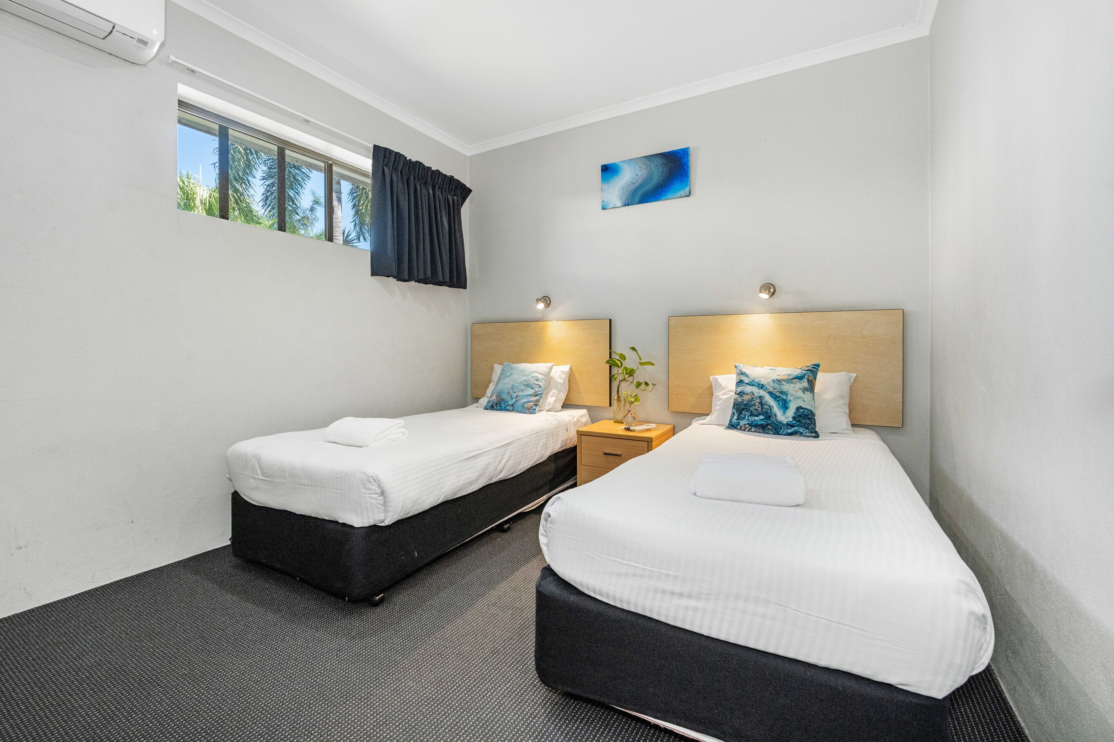 A hotel room with two beds, a window with curtains, a small bedside table with a lamp, and a wall-mounted painting.