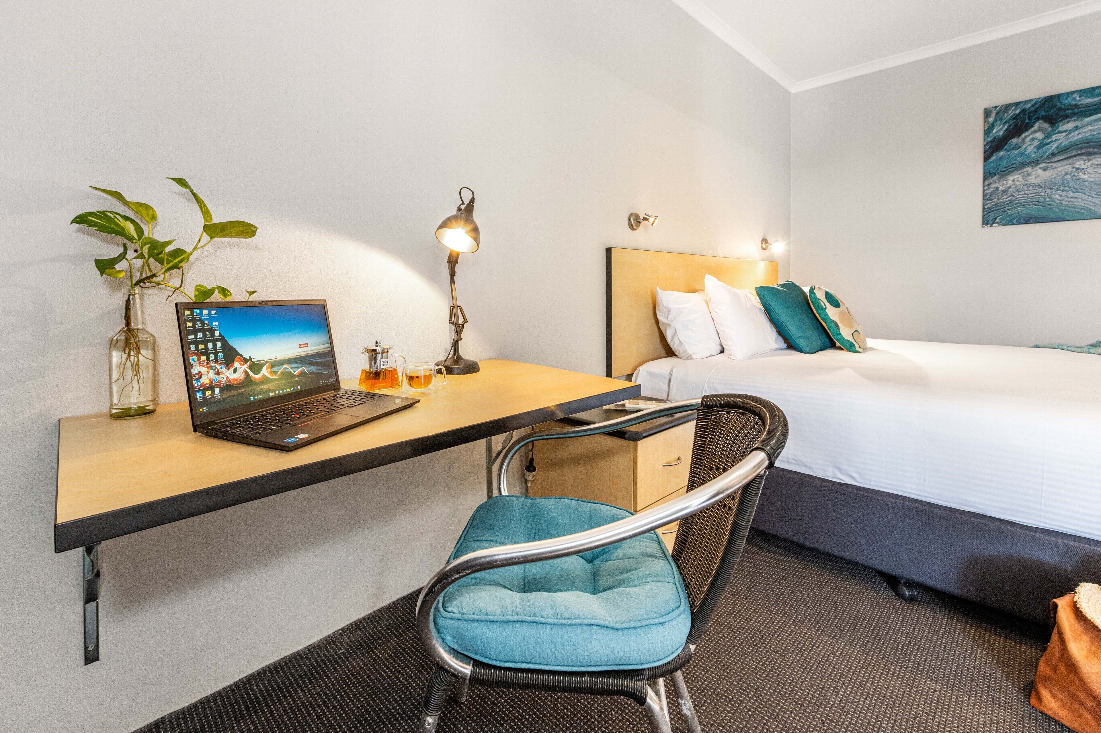 A hotel room with a bed, a desk with a laptop, a chair, a plant, and a painting on the wall.