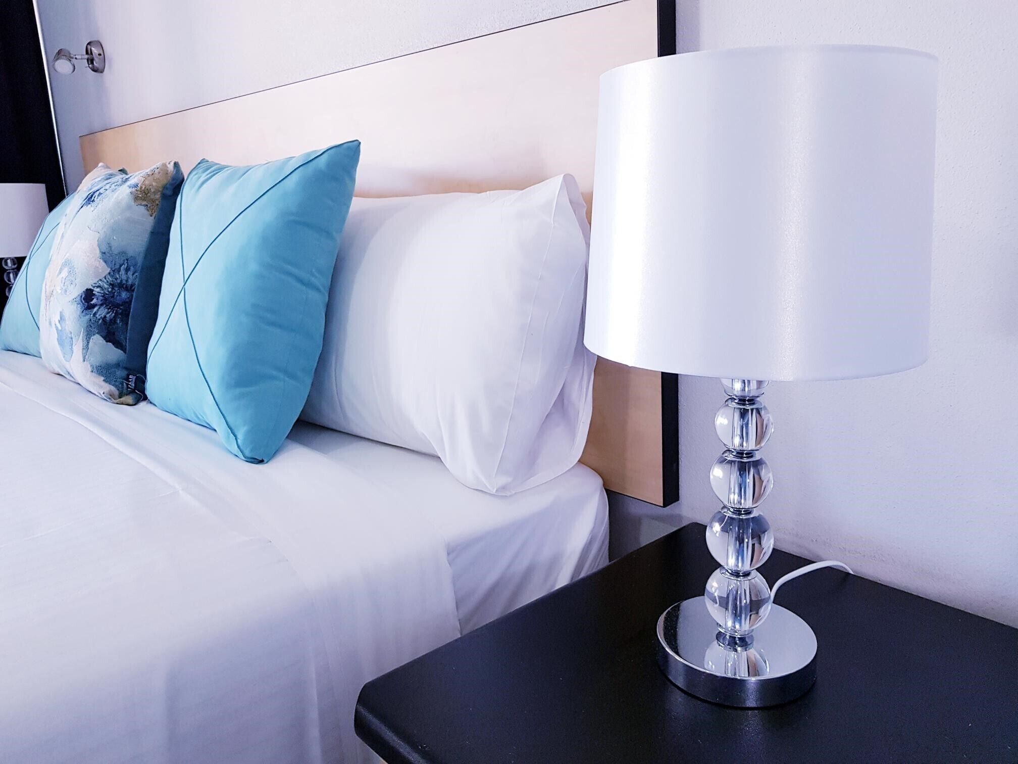A neatly made bed with white and blue pillows, a white headboard, and a modern table lamp on a black nightstand.