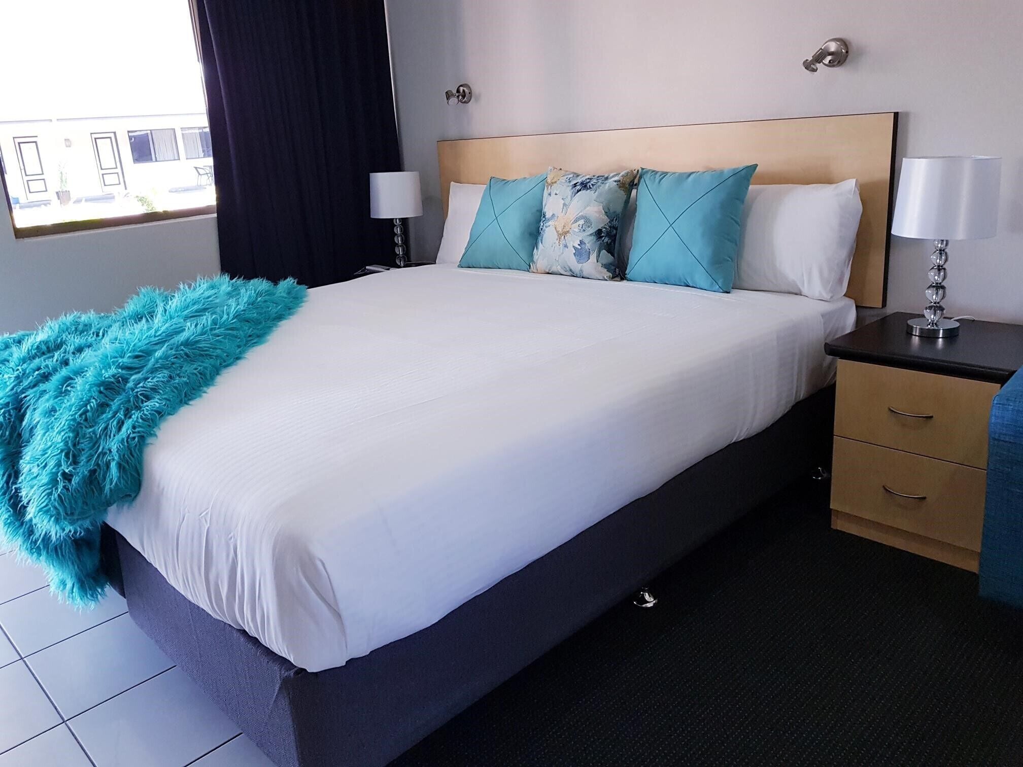 A hotel room with a large bed, a nightstand, a lamp, and a view of the outside.