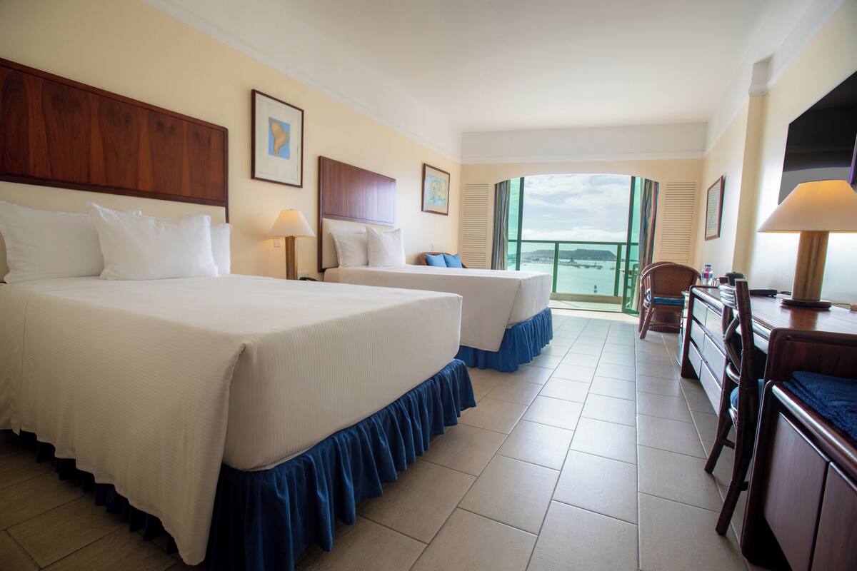 A hotel room with two beds, a desk, and a view of the sea.