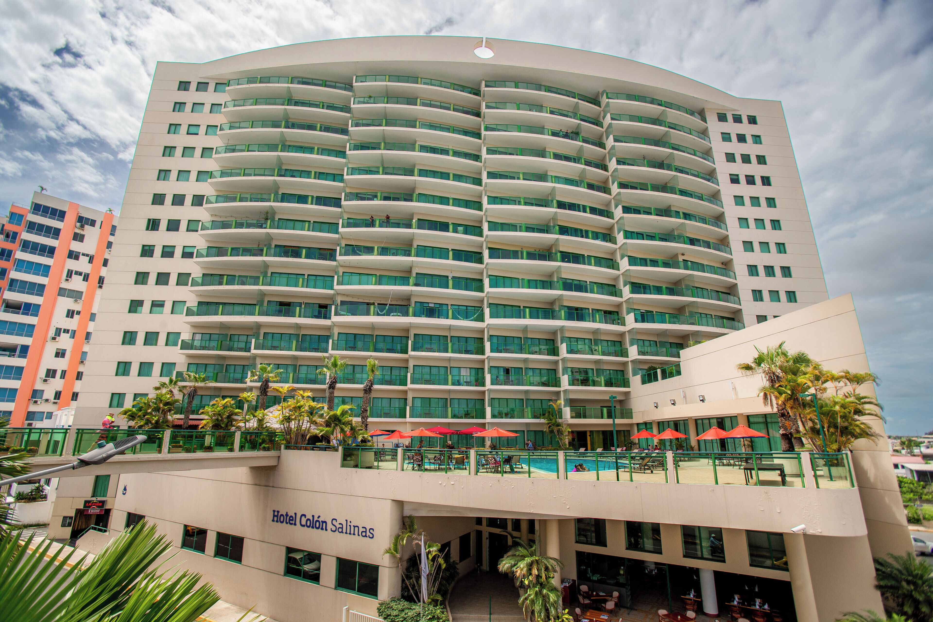 Un hotel de varios pisos con fachada curva, balcones y una zona de piscina con sombrillas.