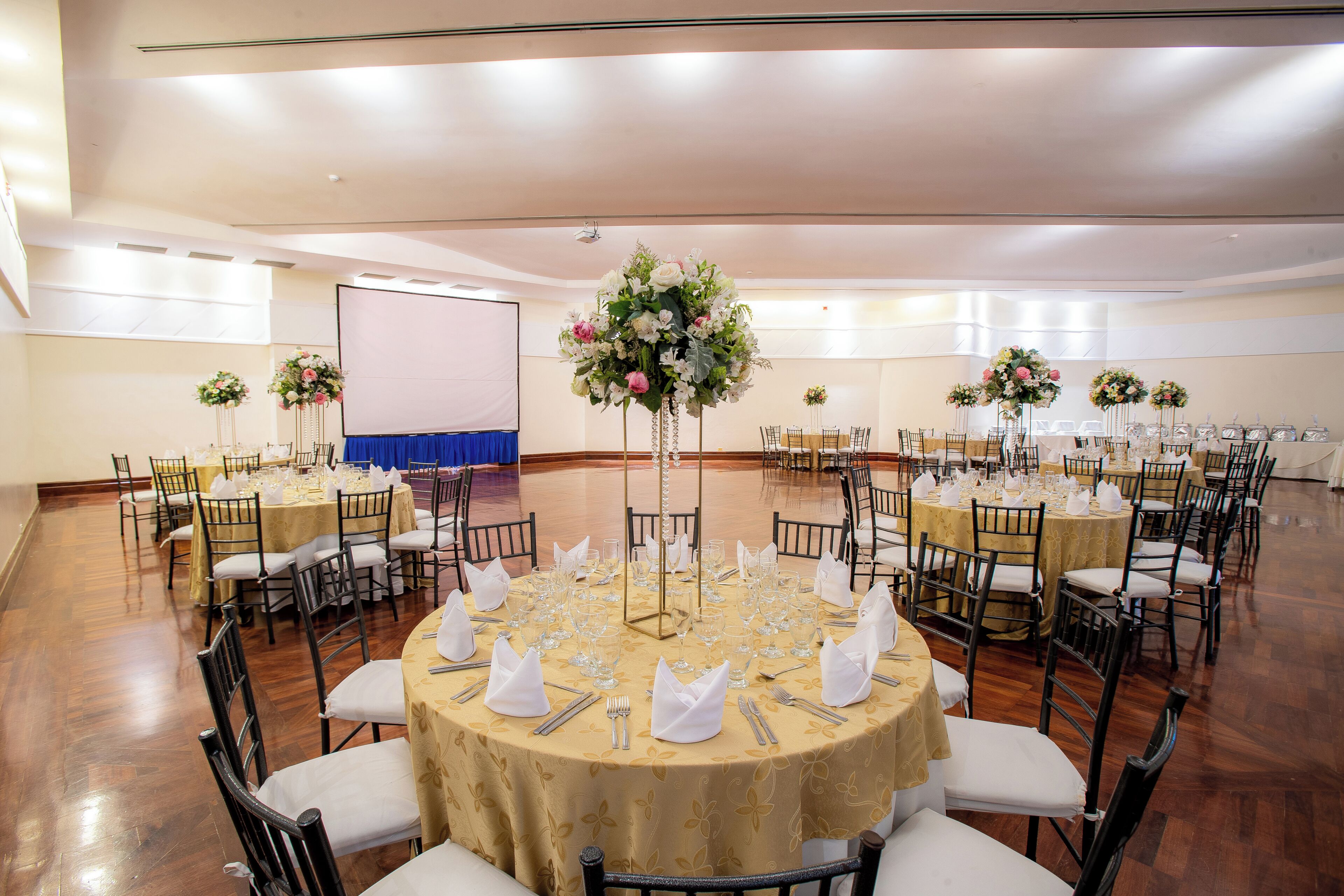 A large banquet hall with round tables set for a formal event, featuring floral centerpieces and white tablecloths.