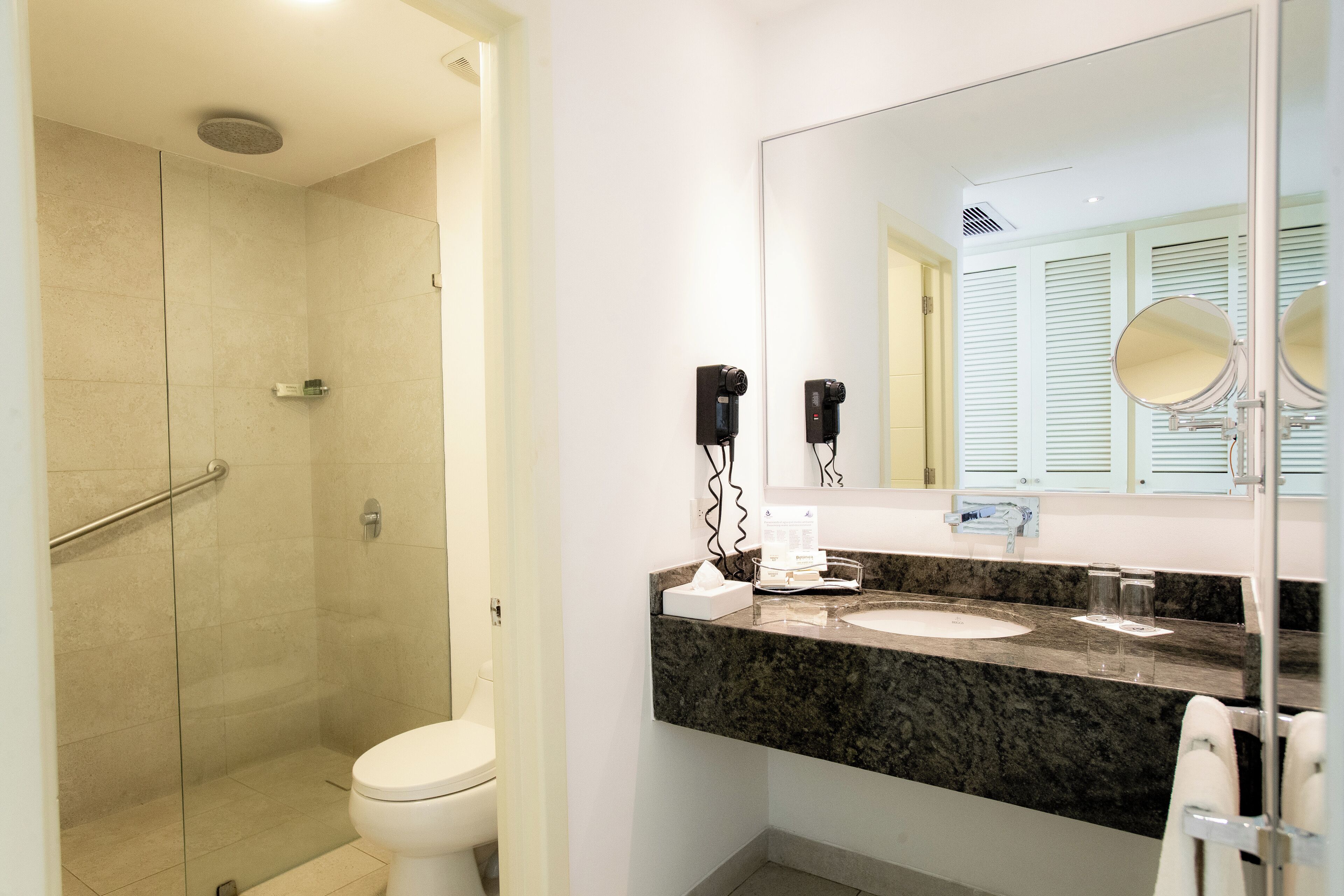 Un baño moderno con ducha acristalada, inodoro blanco, encimera de granito, espejo grande y toallero.