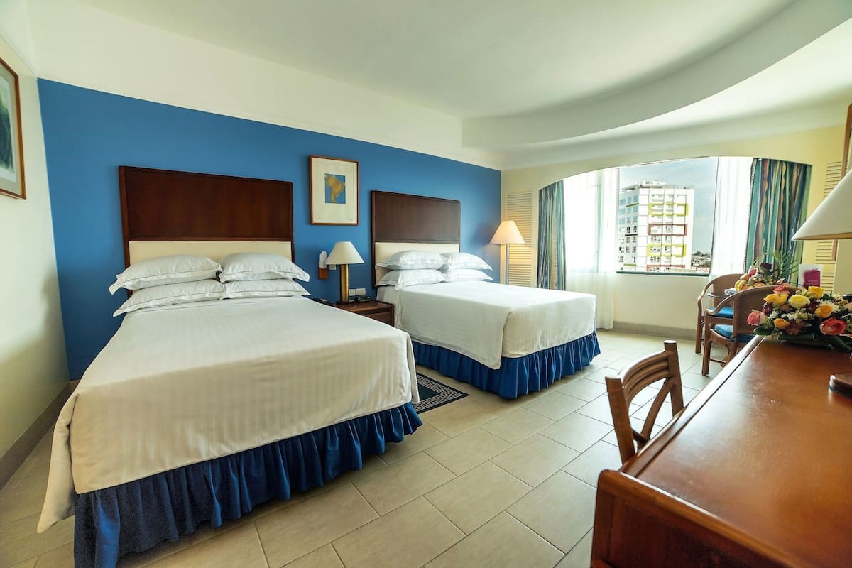 A hotel room with two beds, a dining table, a window with curtains, and a view of buildings outside.