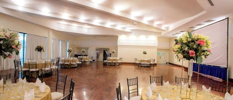 A large banquet hall with multiple round tables set for dining, each with white tablecloths and neatly arranged place settings.