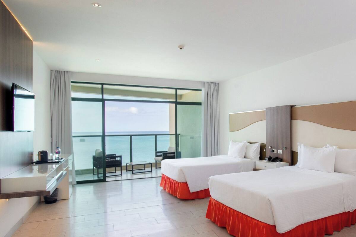 A hotel room with two beds, a desk, a TV, and a balcony with a view of the sea.