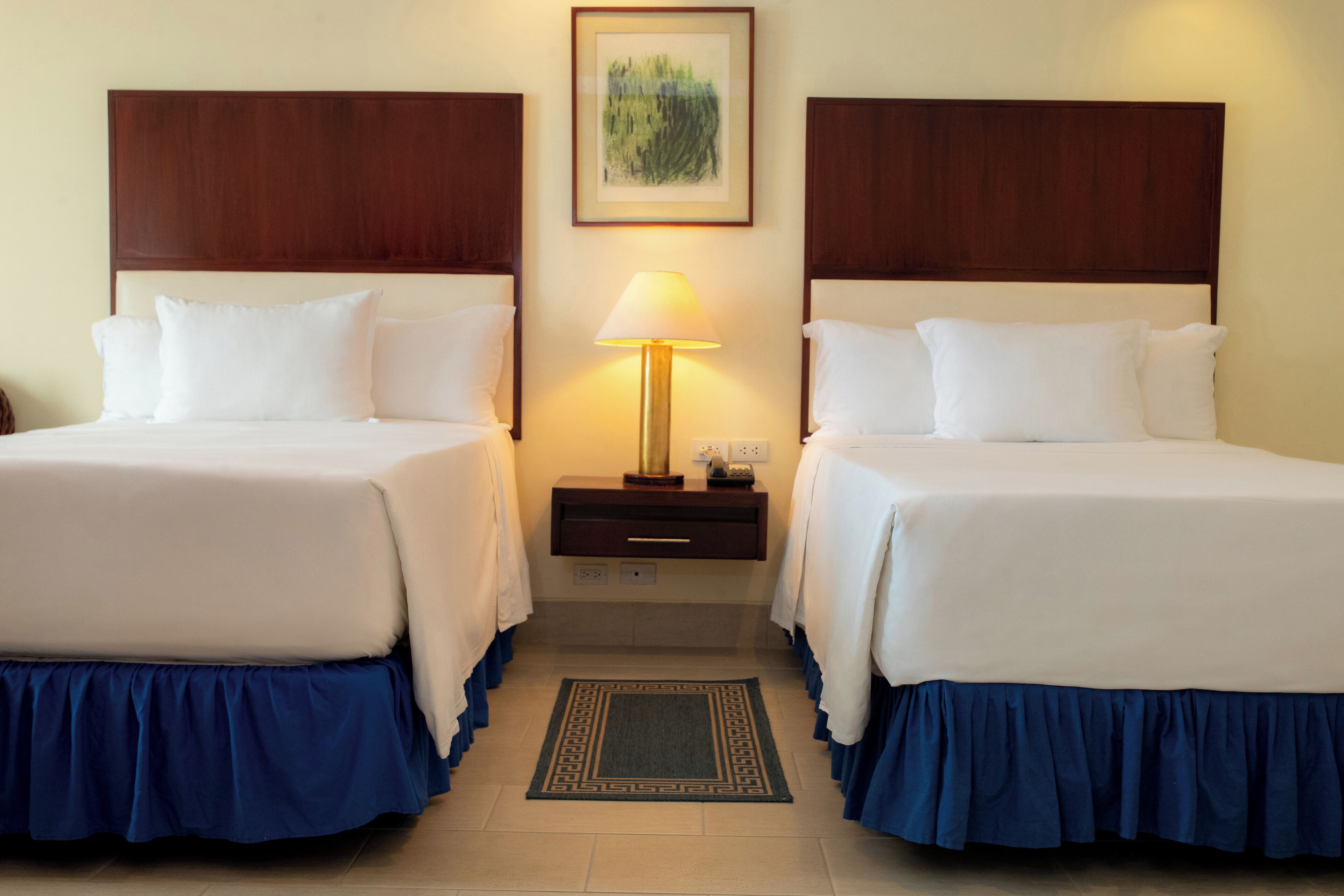 A hotel room with two beds, a nightstand with a lamp, and a framed picture on the wall.