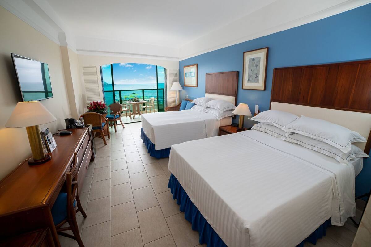 A hotel room with two beds, a desk, a chair, a table, and a view of the ocean.