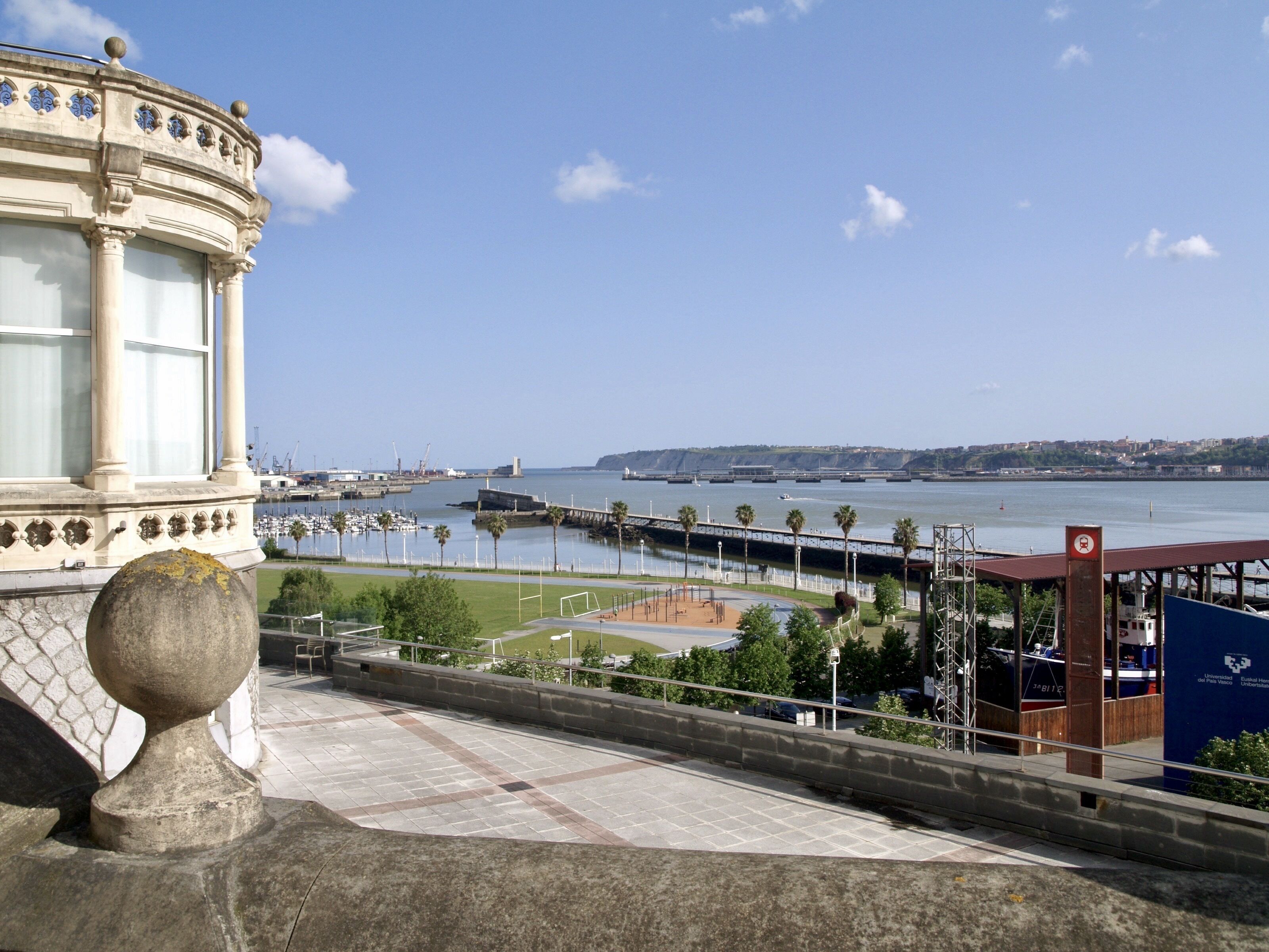 Vista costanera amb un edifici històric, un port esportiu i un cel serè.