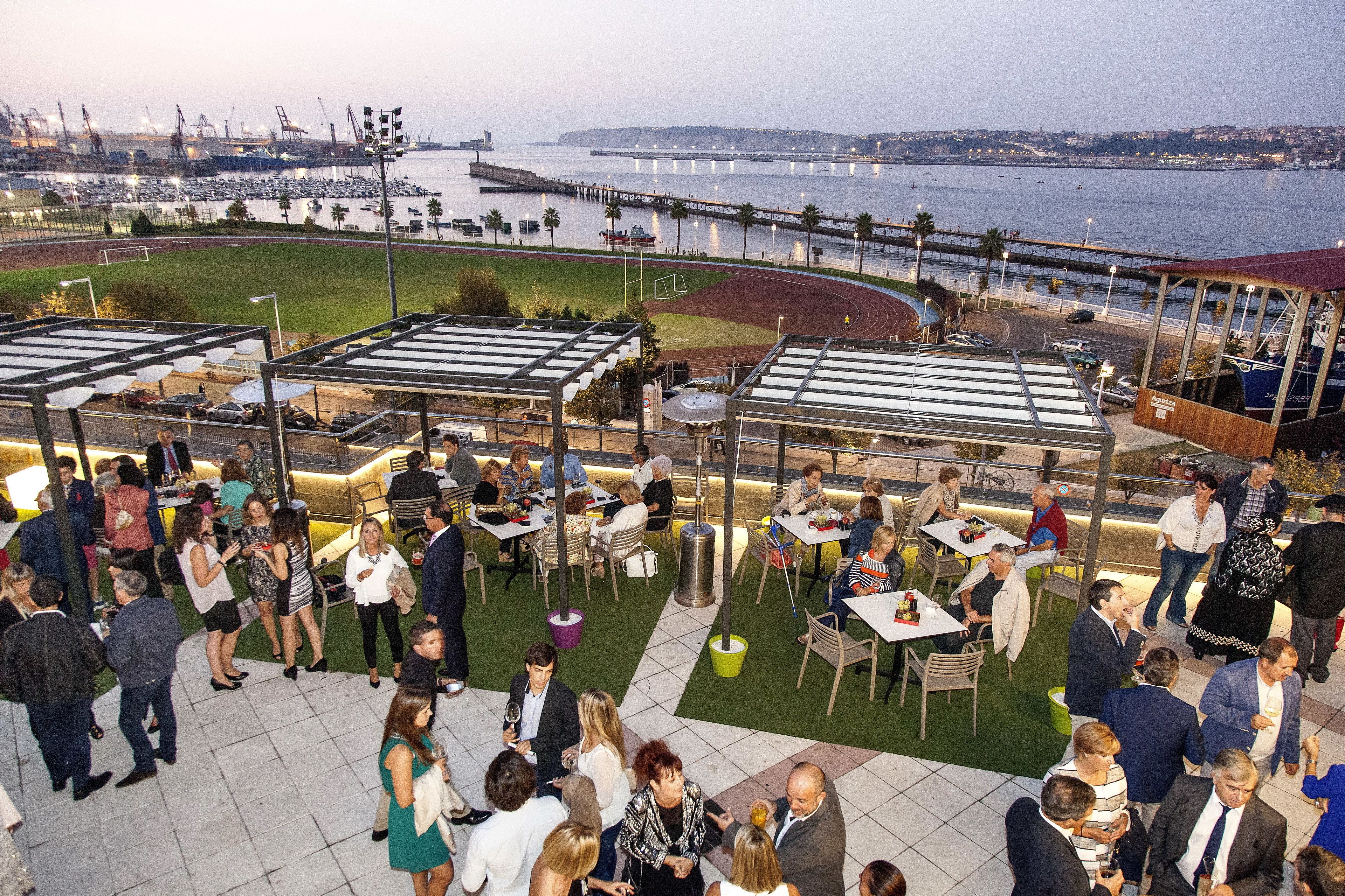 Un patio al aire libre con mesas y sillas, vista al puerto deportivo y un campo de deportes al fondo.