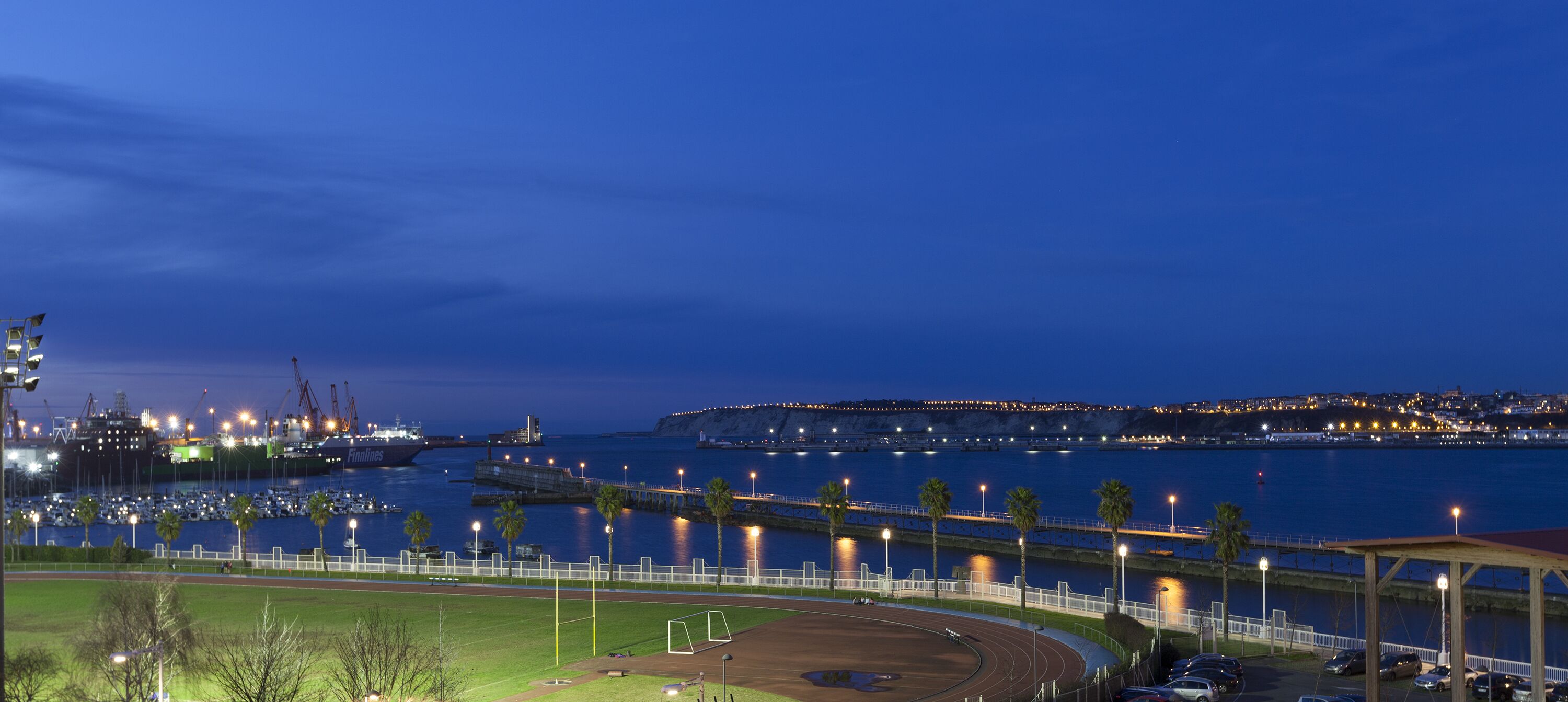 Vista costera con un puerto deportivo, un campo de deportes y un paisaje urbano distante al atardecer.