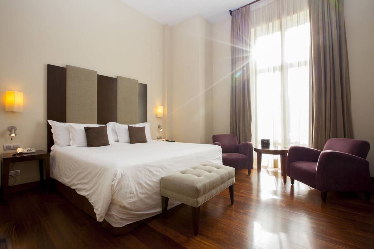 Ein Hotelzimmer mit einem großen Bett, zwei Sesseln, einem kleinen Tisch und einem Fenster mit Vorhängen.