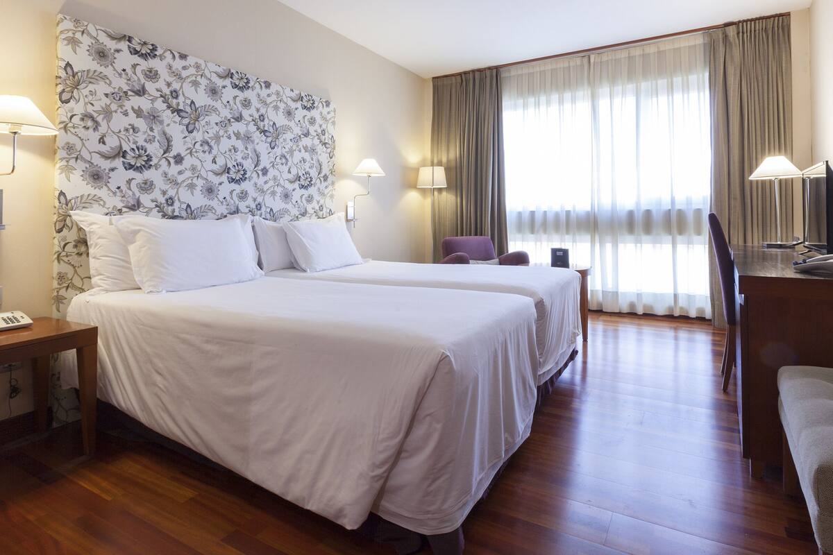Ein Hotelzimmer mit zwei Betten, einem Schreibtisch, einem Stuhl und einem großen Fenster mit Vorhängen.