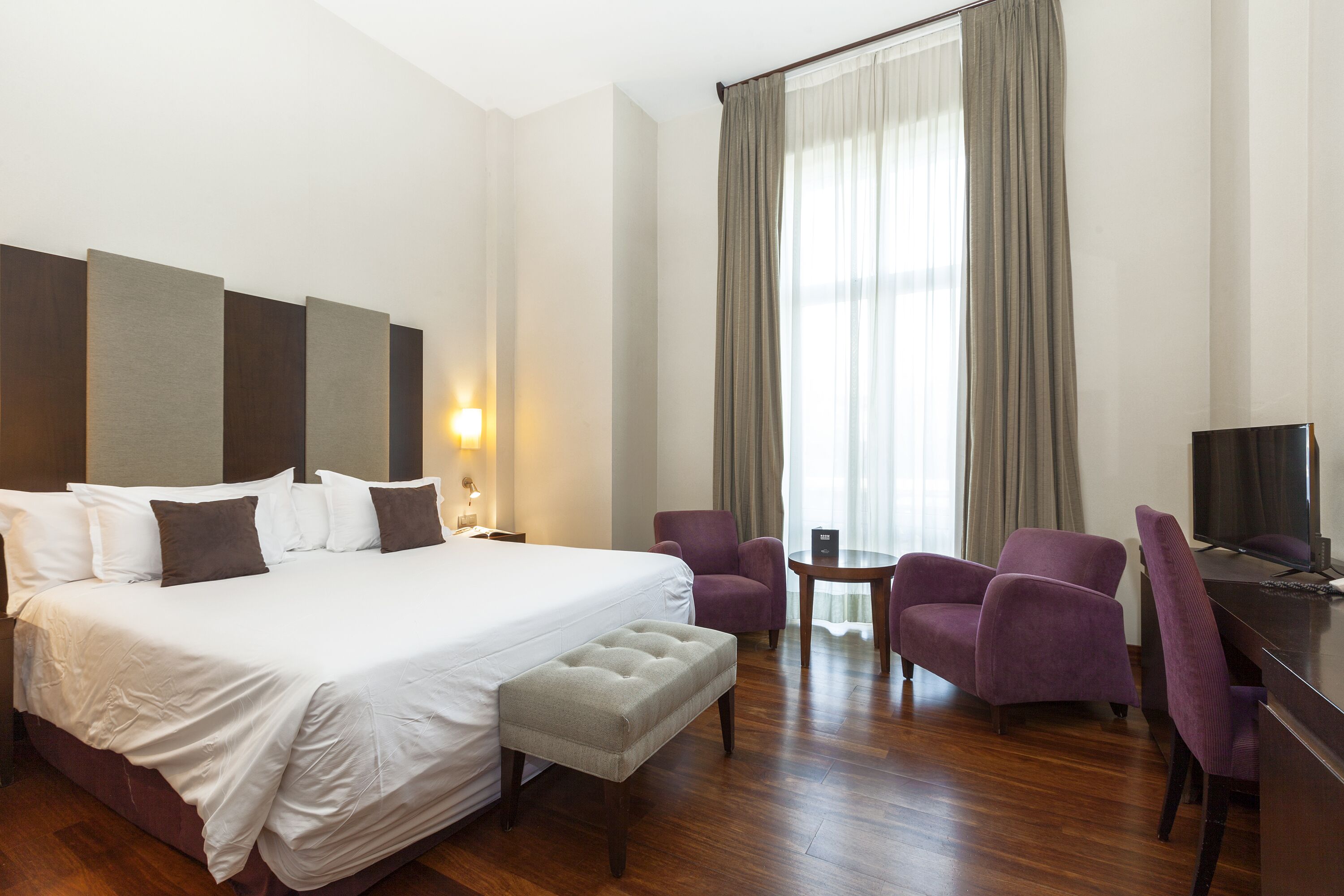 A hotel room with a large bed, two armchairs, a small table, and a desk with a TV.