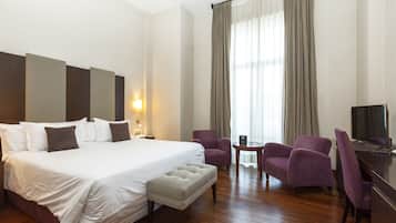 A hotel room with a large bed, two armchairs, a small table, and a desk with a TV.