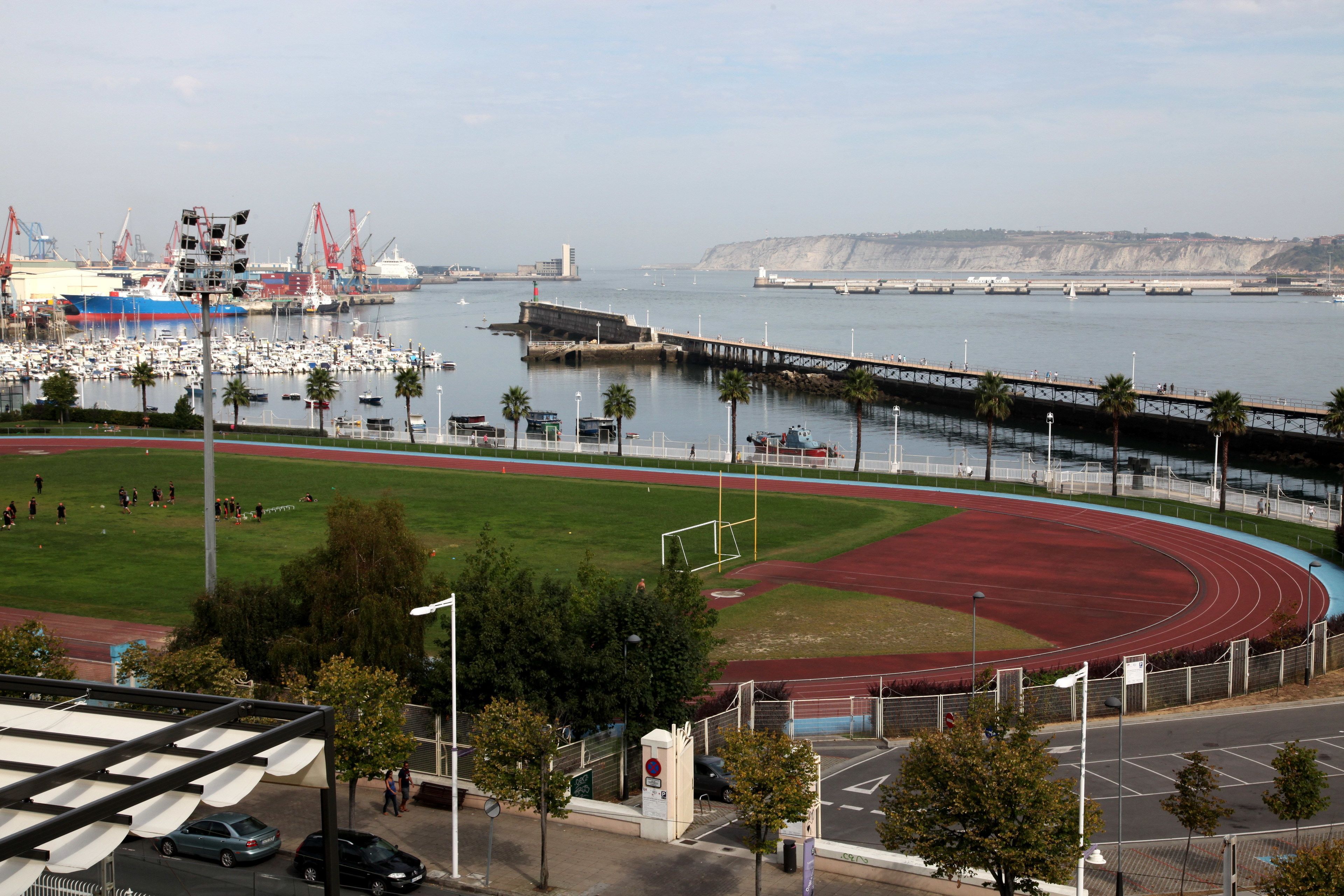 Un complejo deportivo costero con pista de atletismo, campo de fútbol y un puerto deportivo con barcos.