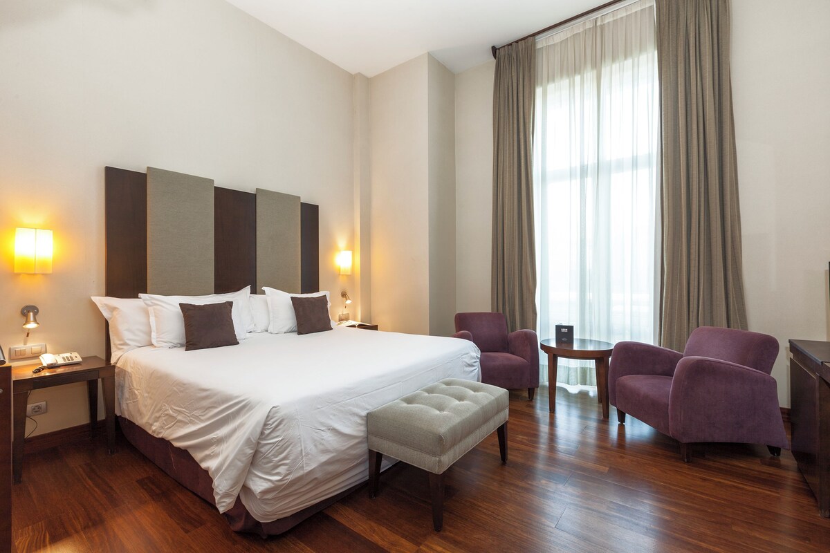 Habitación de hotel con una cama grande, dos mesitas de noche, un escritorio, una silla, una pequeña mesa y un ventanal con cortinas.